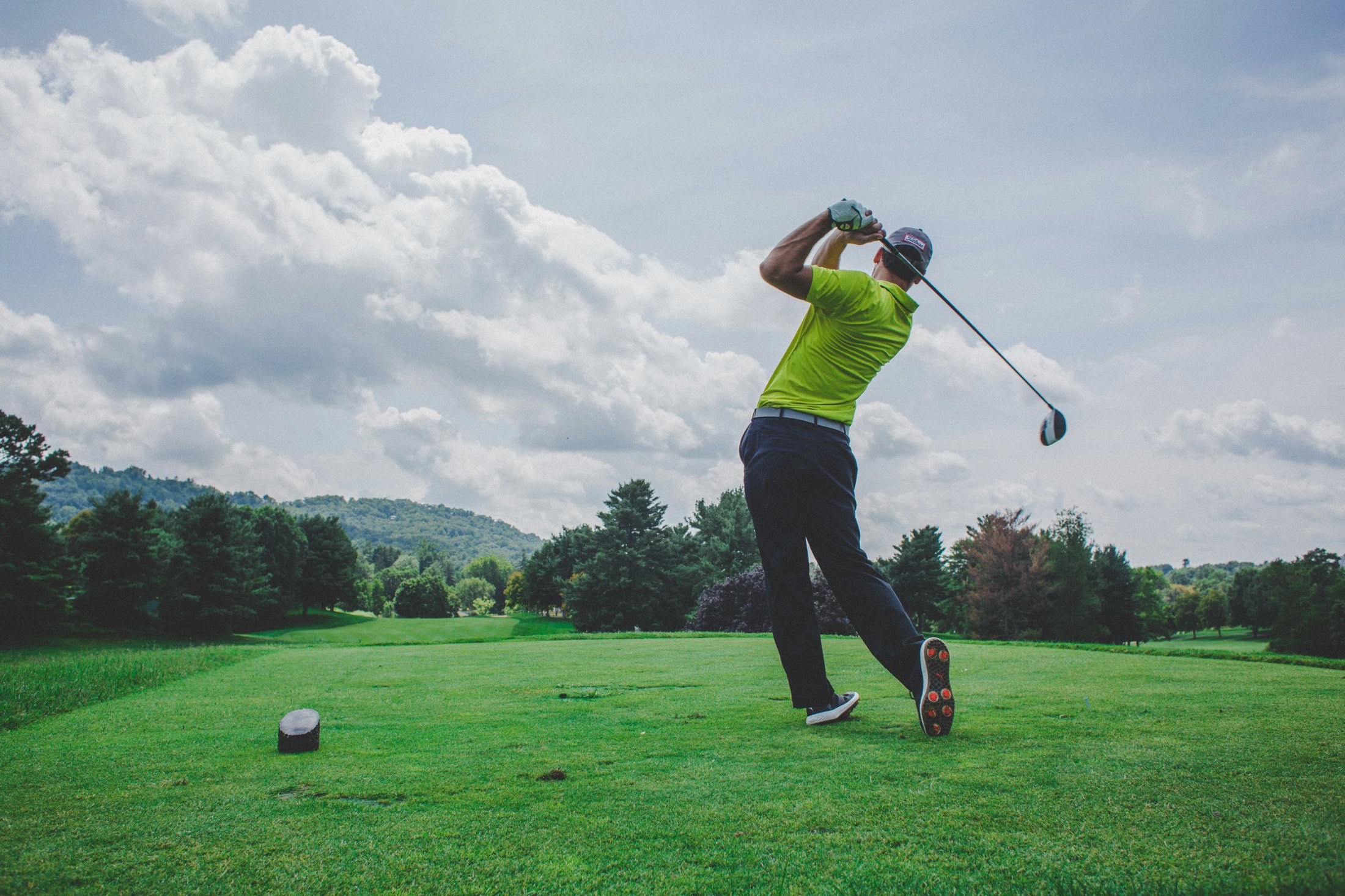 A golfer following through on a tee shot on a fairway framed by trees and hills