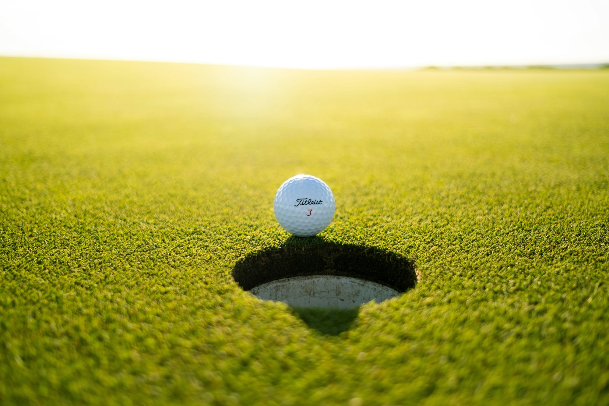A golf ball resting on the very lip of the hole, backlit by low sun